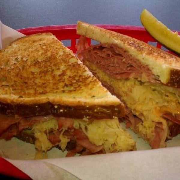 A grilled Reuben sandwich, cut in half, with corned beef, sauerkraut, and melted cheese, served with a pickle spear.