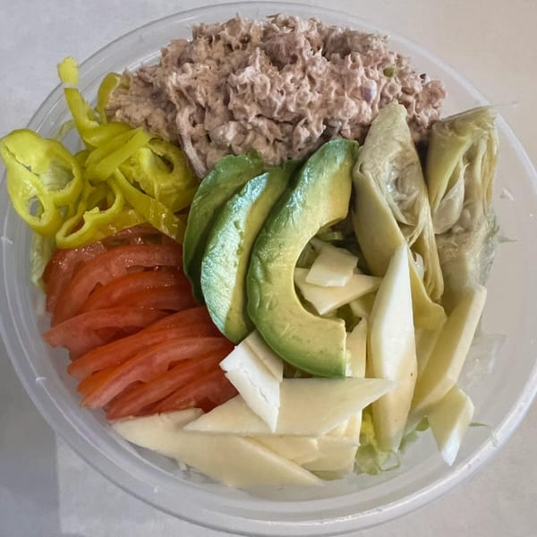 A tuna salad with sliced tomatoes, avocado, banana peppers, artichoke hearts, and cheese in a clear bowl.